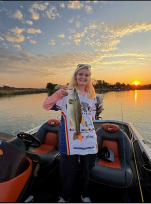 Person on a boat holding a large fish at sunset. The boat has red and black seats, and the water reflects the orange and pink hues of the sky as the sun sets on the horizon.