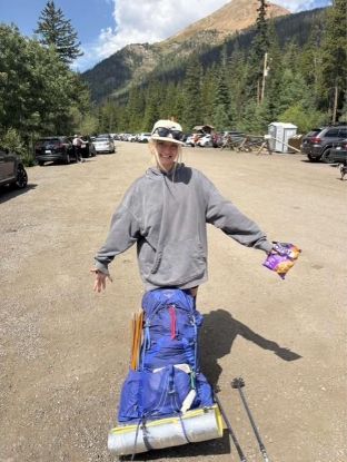 Person standing on a dirt parking area surrounded by cars and trees, wearing a gray hoodie and holding a snack bag. A large blue hiking backpack with gear and a rolled sleeping pad is on the ground in front, with trekking poles nearby. Mountains and a partly cloudy sky are visible in the background.