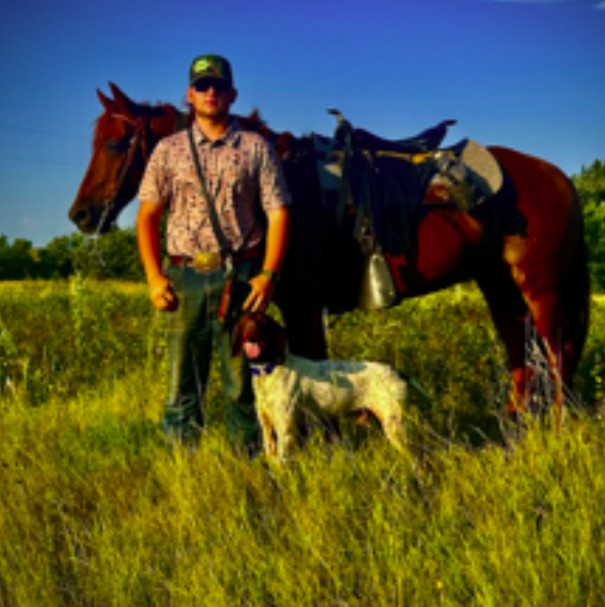 Person standing in a grassy field next to a saddled horse and a dog.