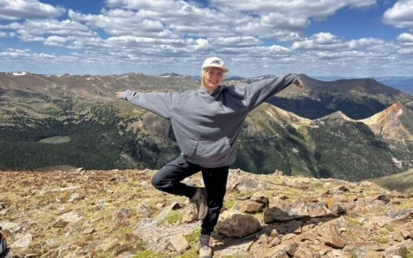 Person standing on a rocky mountain summit in a yoga tree pose, wearing a gray hoodie and cap. The background shows expansive mountain ranges under a bright blue sky with scattered clouds.