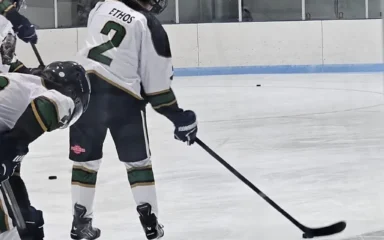 Ice hockey players skate during a game, with one player wearing a white jersey marked “ETHOS” and number 2 controlling the puck on the ice near the boards while another player moves alongside.