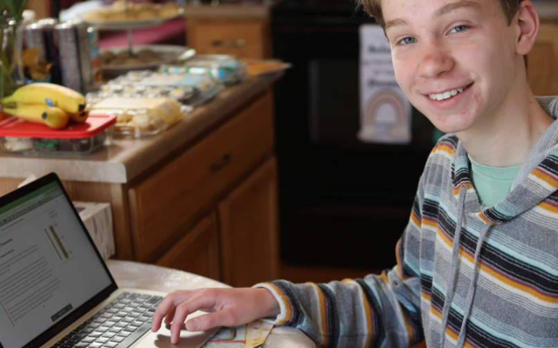 A person sitting at a kitchen table using a laptop, with a notebook and pen nearby and a small cat resting on their lap.