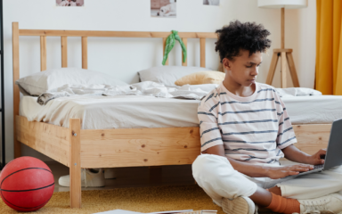A student sitting on the floor in a bedroom, using a laptop near a bed, with notebooks and a basketball nearby.