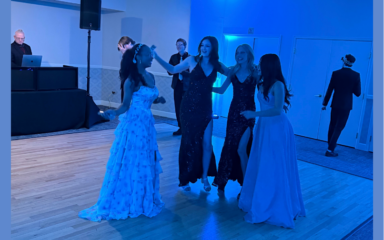 Four people in formal evening attire dance together on a wooden floor under blue lighting at an indoor event, while additional guests and a DJ setup are visible in the background near a wall and door.