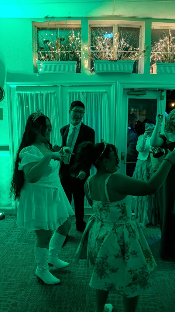 A group of people in formal attire dance together inside a venue lit with green lighting. In the foreground, two individuals wearing tiaras and dresses move with their arms raised, while another person in a suit stands nearby. In the background, decorative window boxes filled with branches and string lights hang above curtains, and additional guests stand near a doorway taking photos.